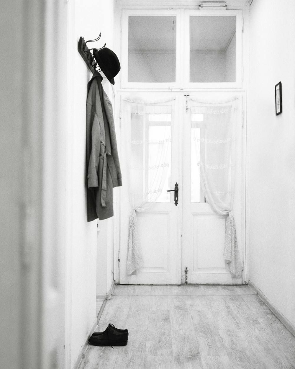 Dans un couloir blanc, en noir et blanc, un manteau et un chapeau melon pendent à une rangée de crochets sur le mur de gauche ; une porte vitrée occupe le fond et une paire de chaussures est posée au sol.