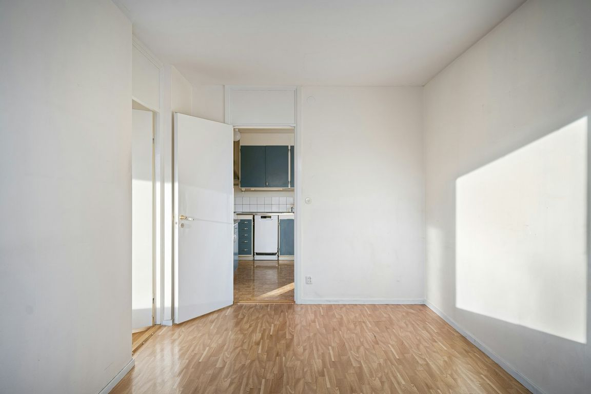 Une pièce vide aux murs blancs et au parquet clair, avec une porte ouverte sur une cuisine et un rayon de soleil sur le mur de droite.