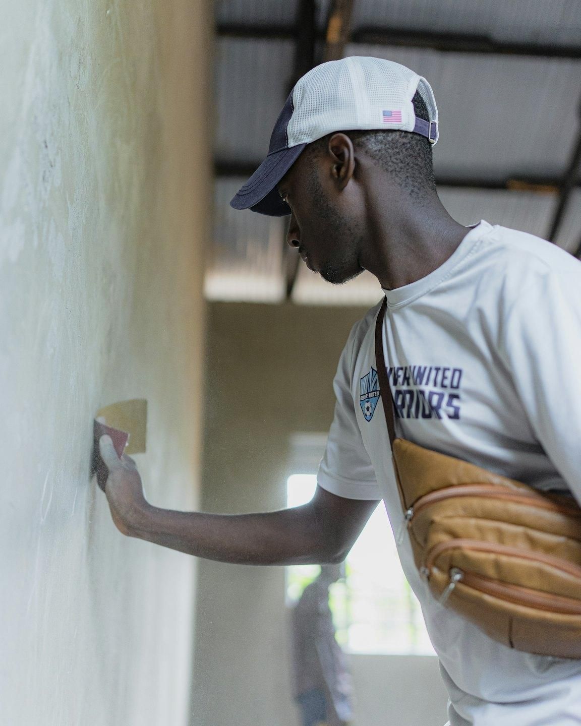 Un homme en casquette ponce un mur à la main avec un morceau de papier abrasif.