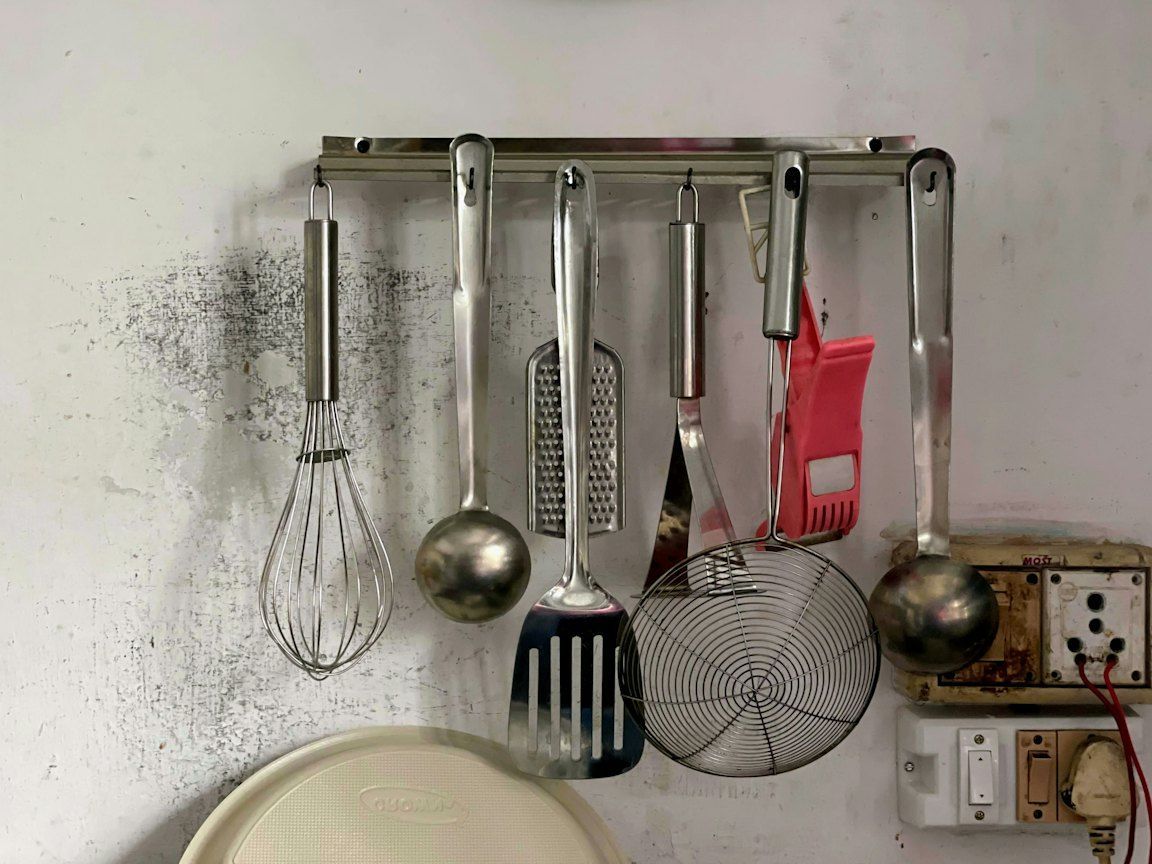 Un fouet, deux louches, une râpe, une spatule et une écumoire pendent à une barre métallique à crochets fixée sur un mur clair.