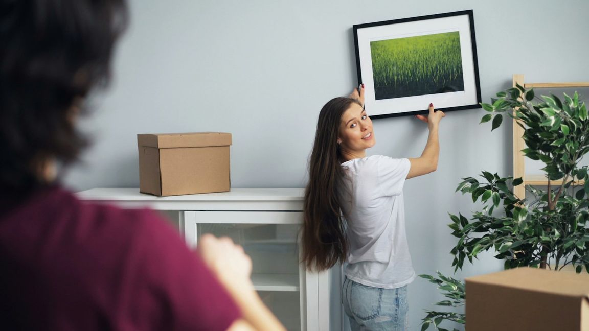 Une jeune femme en t-shirt blanc pose un cadre noir à passe-partout blanc sur un mur gris clair, entourée de cartons de déménagement.