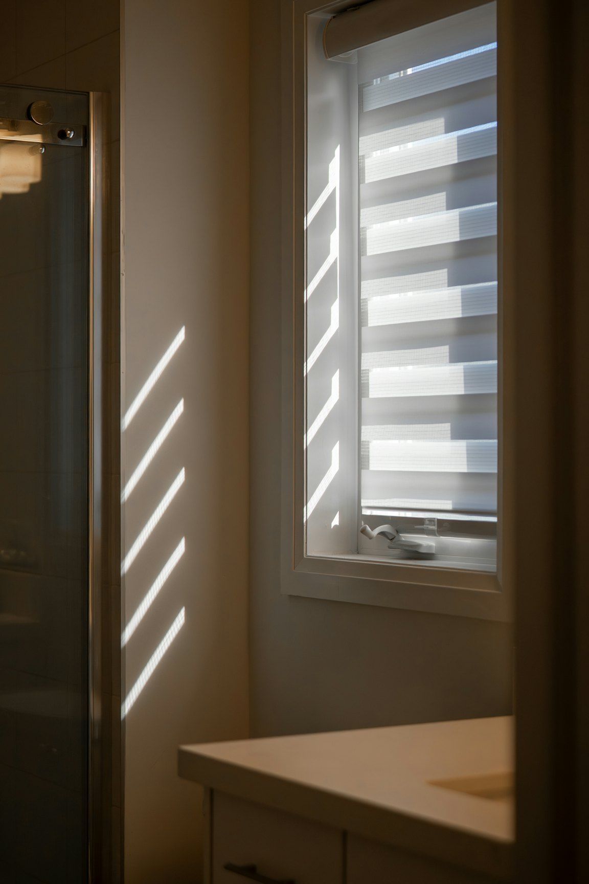 Un store à bandes alternées translucides et opaques devant une petite fenêtre, à côté d’une paroi de douche, avec des rayures de soleil sur le mur.